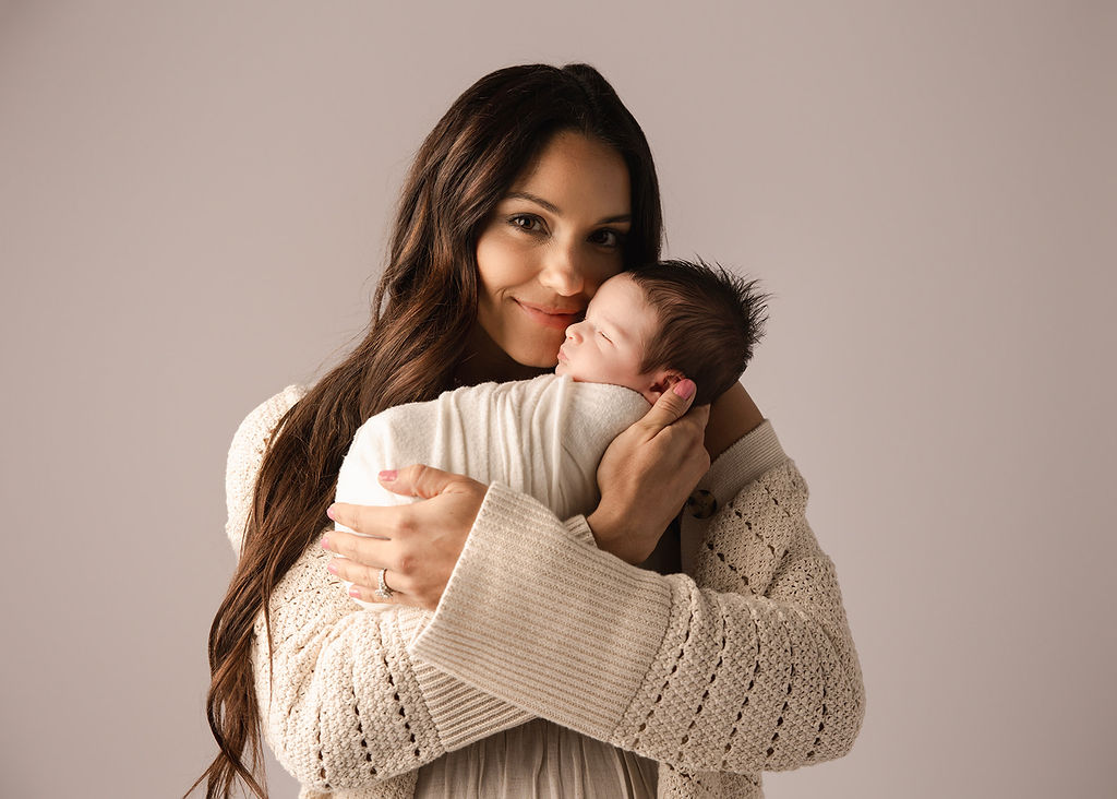 A smiling new mother stands cradling her sleeping newborn baby against her cheek in a studio