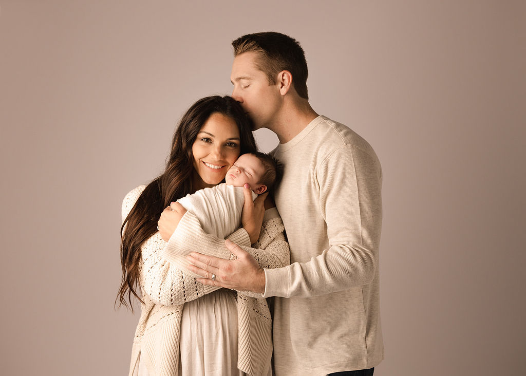 A new father kisses his wife's head as she smiles with her newborn baby against her cheek in a studio after meeting a nanny in Boise