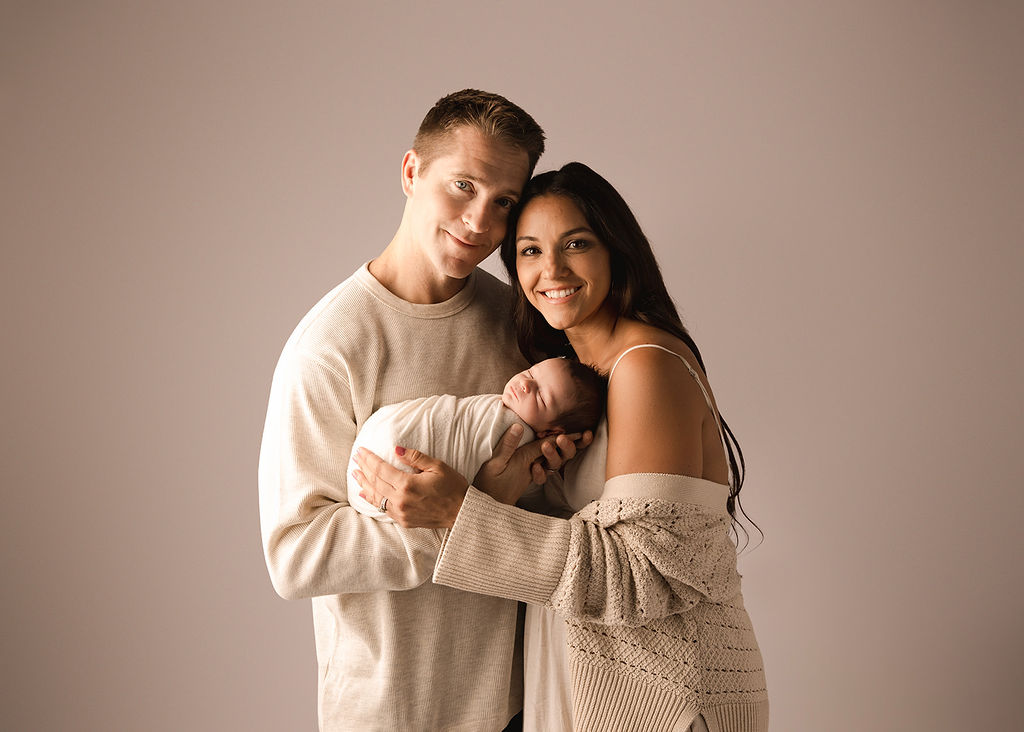 Happy new parents stand in in a studio holding their sleeping newborn between them after meeting a nanny in Boise