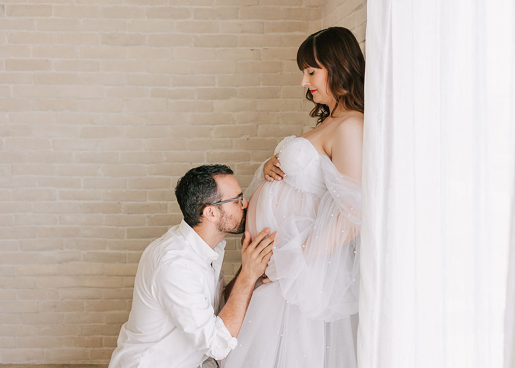 A father to be kisses his wife's bump as she leans on a brick wall