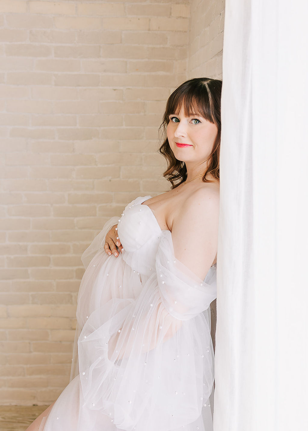 A smiling pregnant woman leans against a window in a studio in a white maternity gown