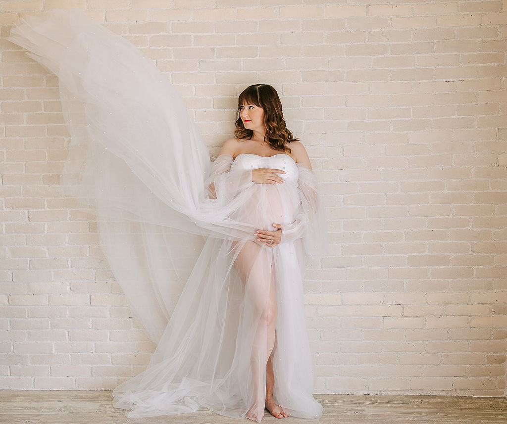 A pregnant woman's maternity gown flows through the air as she leans against a brick wall holding her bump after visiting an OBGYN Boise