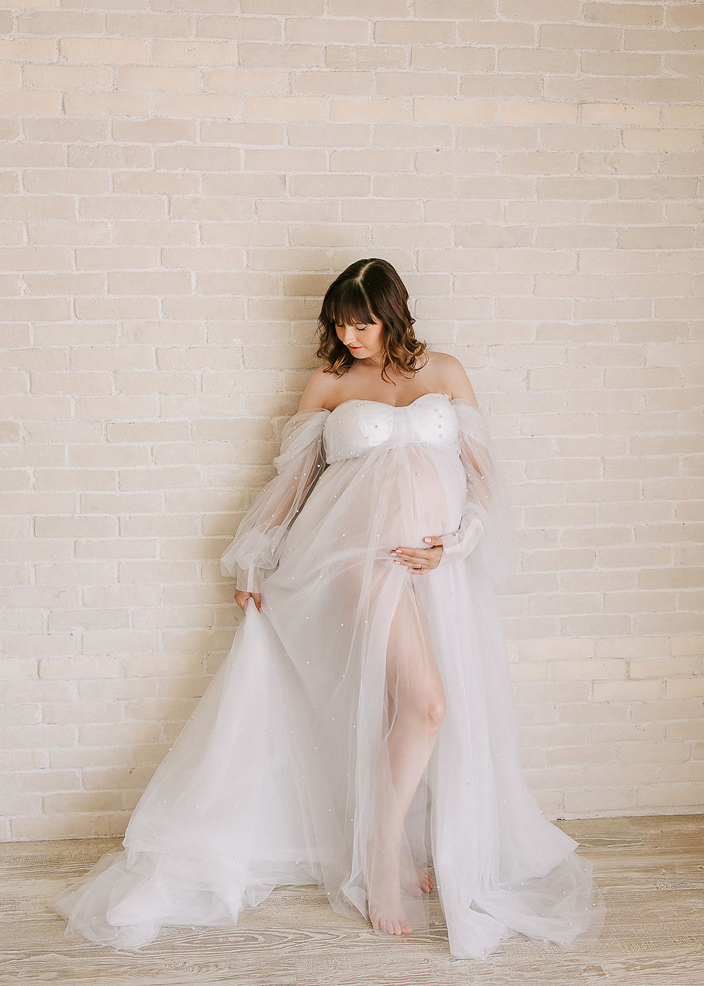 A mother to be plays in her sheer maternity gown while leaning on a brick wall in a studio after visiting an OBGYN Boise