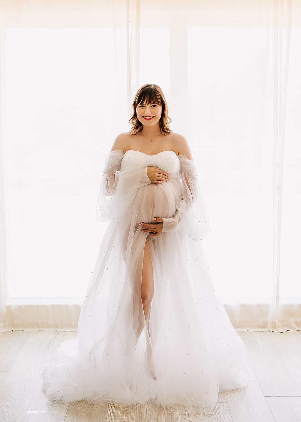 A happy mother stands in a studio window holding her bump in a sheer maternity gown after visiting an OBGYN Boise