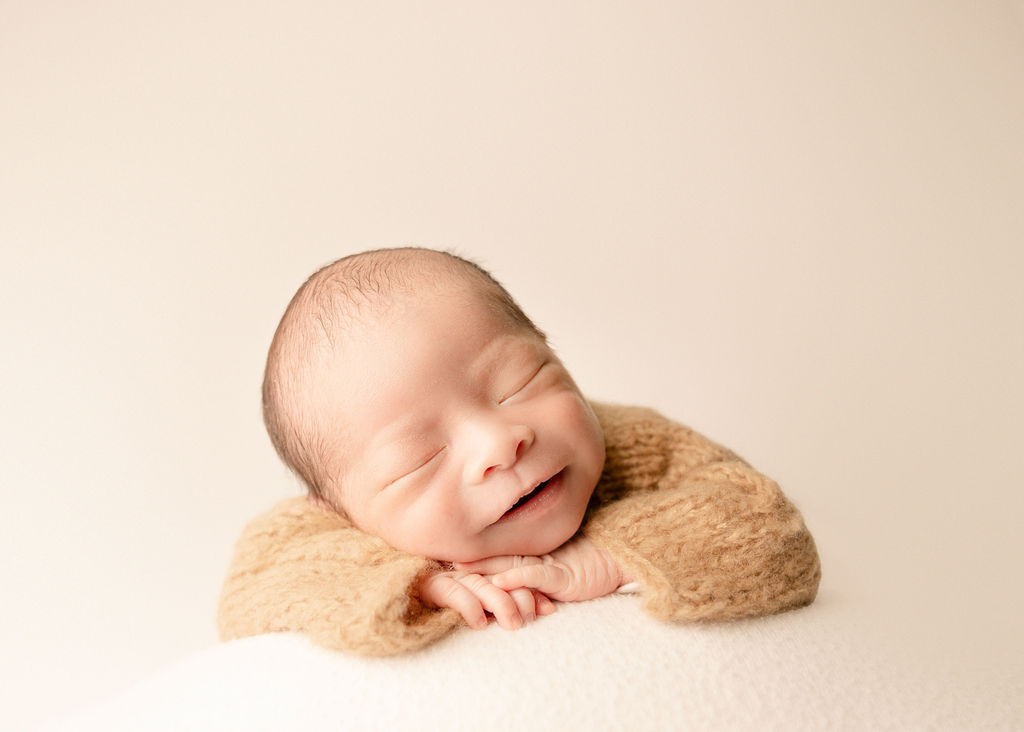 A smiling newborn baby sleeps on its hands in a knit onesie