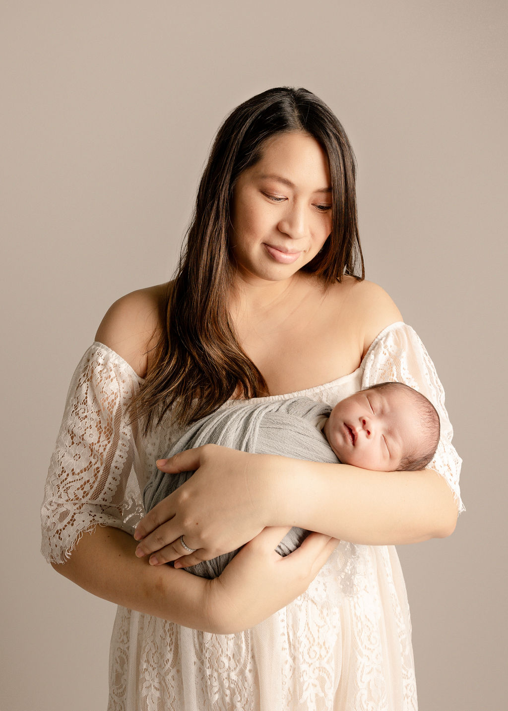 A happy mom smiles down at her sleeping newborn in her arms in a lace dress after visiting a pediatric dentist boise