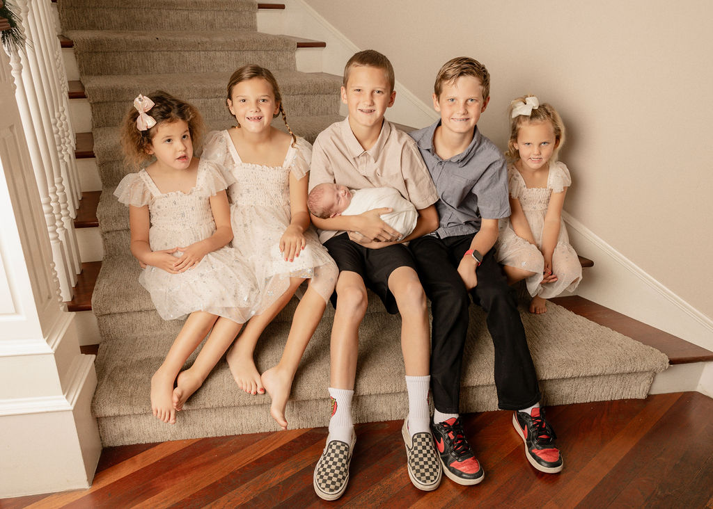 5 young siblings sit on some stairs with the oldest holding a newborn baby sibling