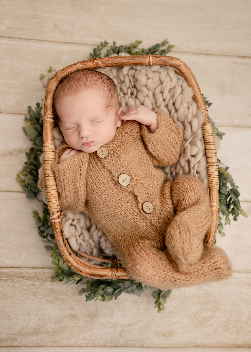 A sleeping newborn baby in a knit brown onesie in a wicker basket in a studio