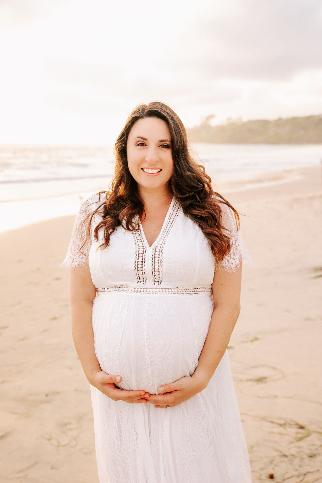 A mom to be in a white lace maternity gown stands on a beach at sunset smiling with hands under her bump