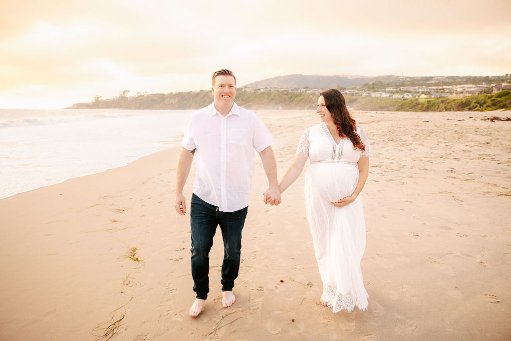 A smiling couple in white holds hands and walks on a beach at sunset while pregnant