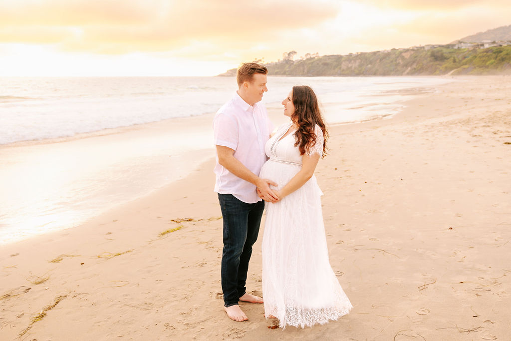 A mom and dad to be hold the bump while standing on an empty beach at sunset