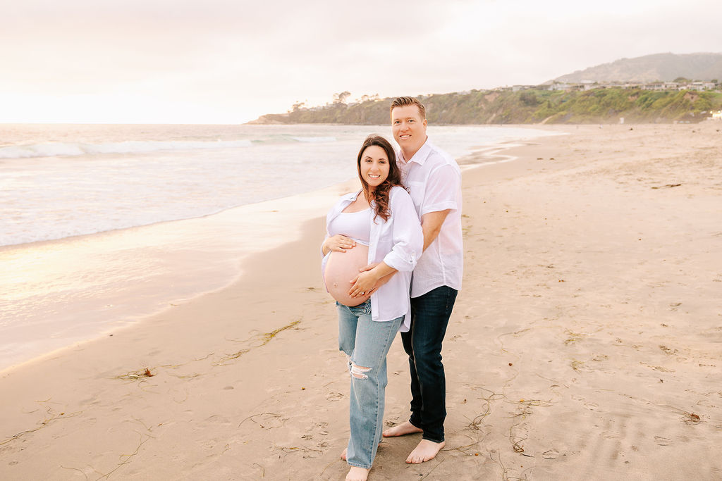 A couple in jeans and white shirts snuggles while standing on a beach at sunset