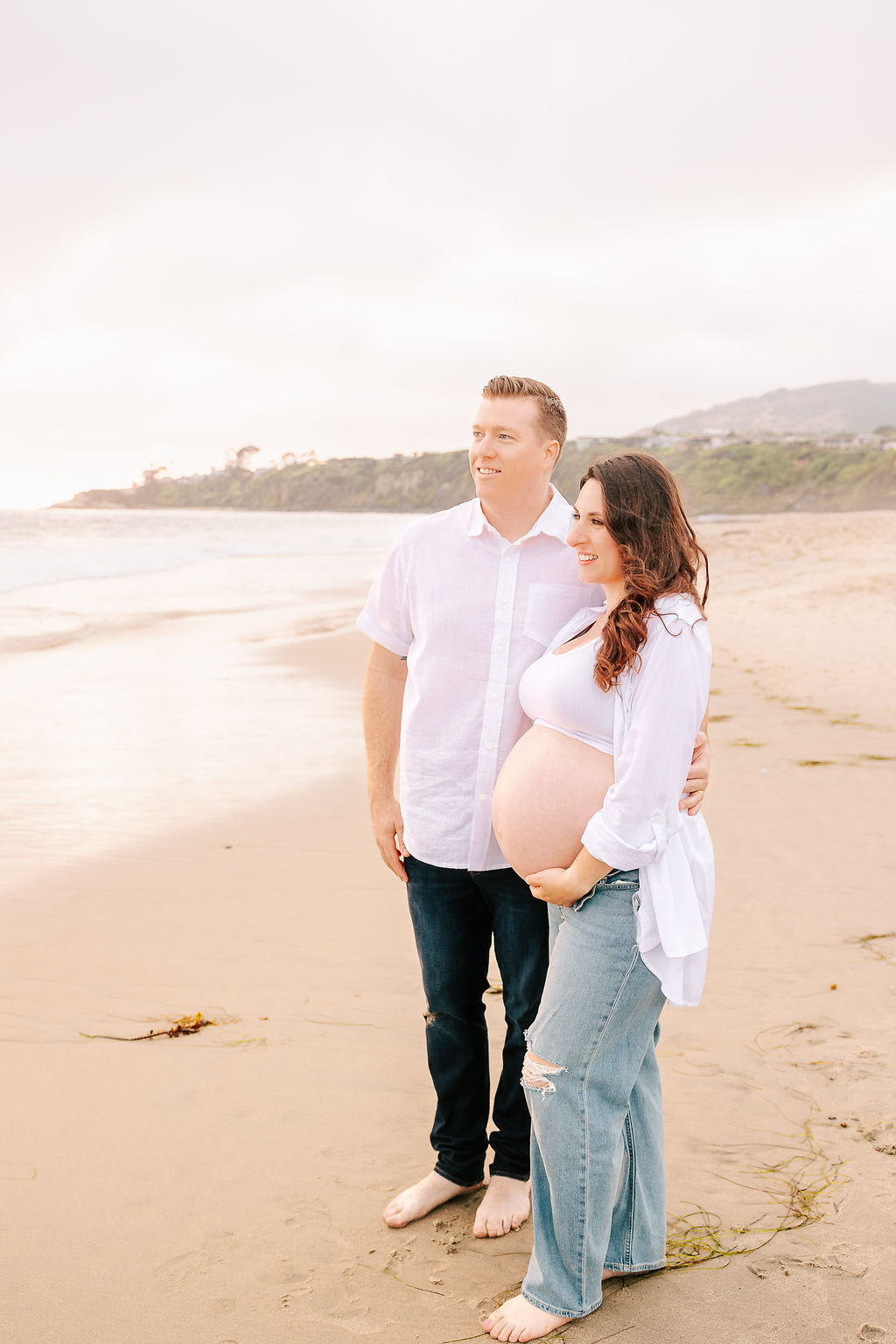 A happy expecting couple stands on a beach admiring the sunset and holding the bump after visiting baby shower venues in Boise, Idaho