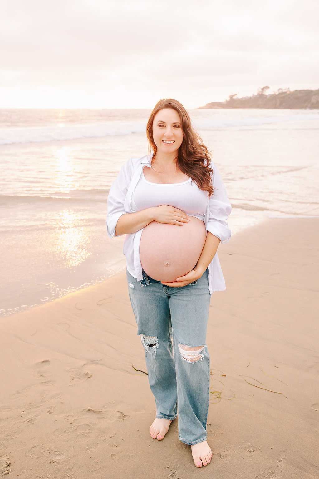 A smiling mother to be walks on a beach in jeans and unbuttoned white shirt at sunset after visiting baby shower venues in Boise, Idaho