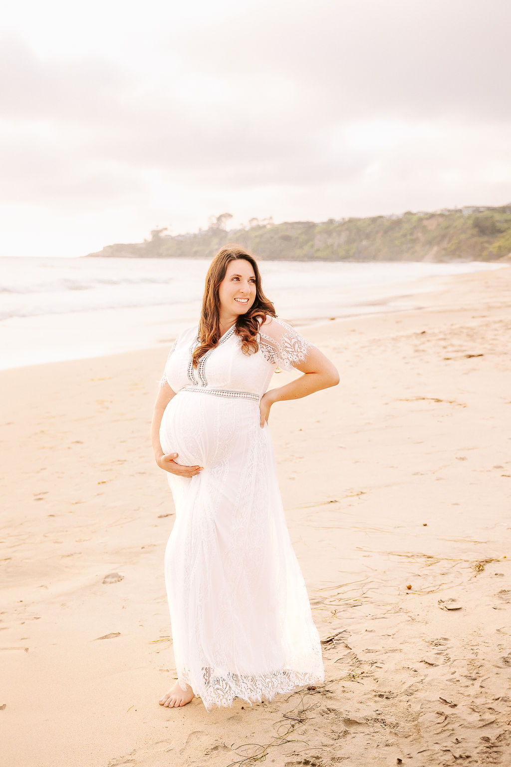 A mother to be in a white maternity gown walks on a beach at sunset with a hand on her back after exploring baby shower venues in Boise, Idaho