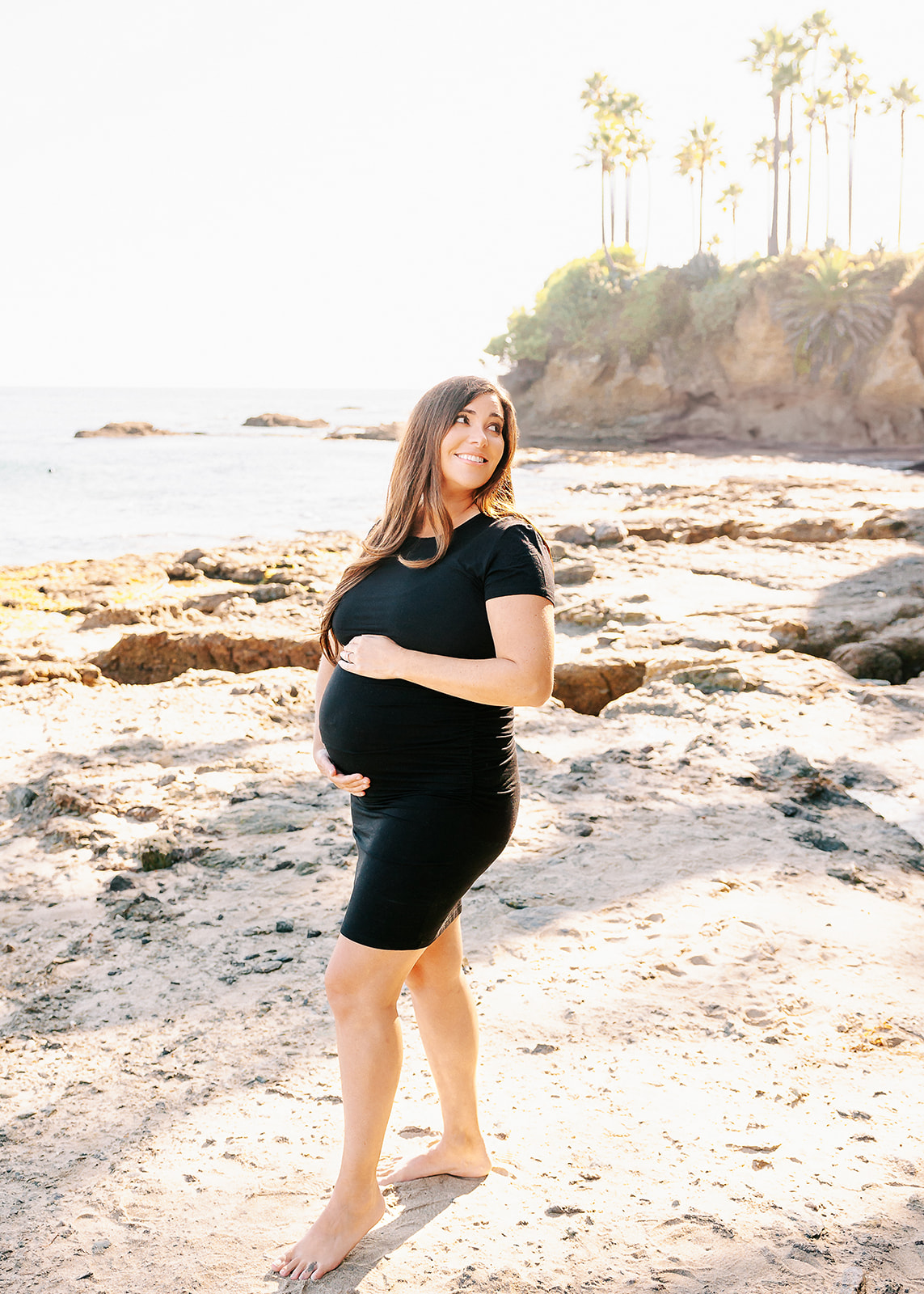 A mom to be smiles over her shoulder while walking on a beach with hands on her bump in a black maternity gown