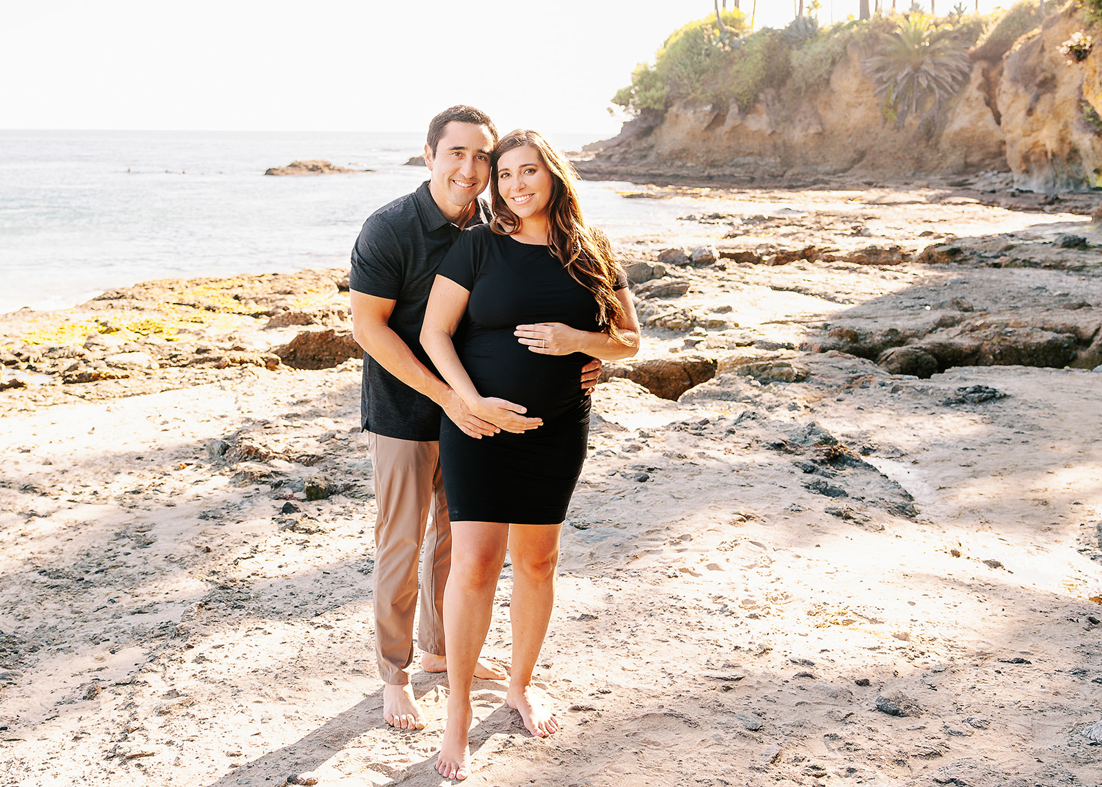 A happy expecting couple in black snuggles while standing on a beach touching heads