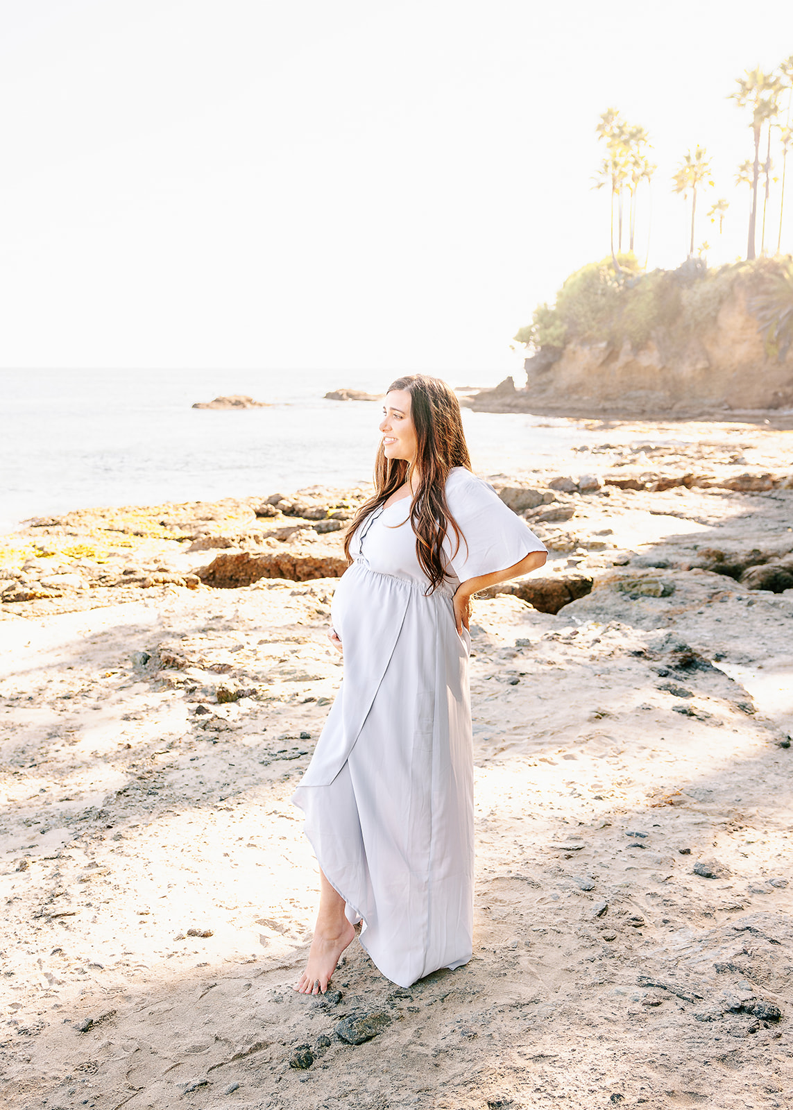 A mother to be in a blue maternity gown rests a hand on her back and bump while standing on a beach at sunset