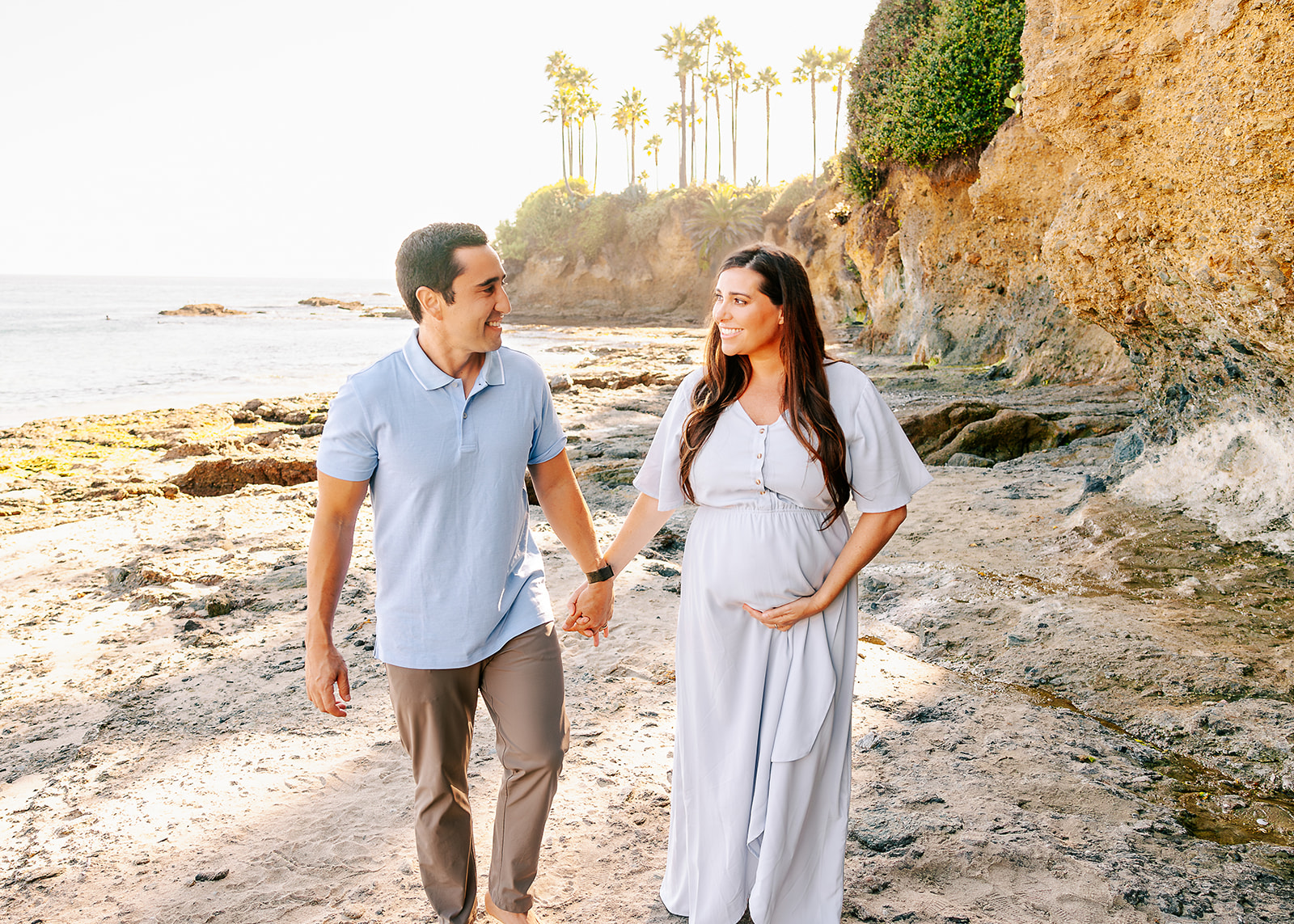 A happy couple walks on a beach holding hands in blue by a large cliff