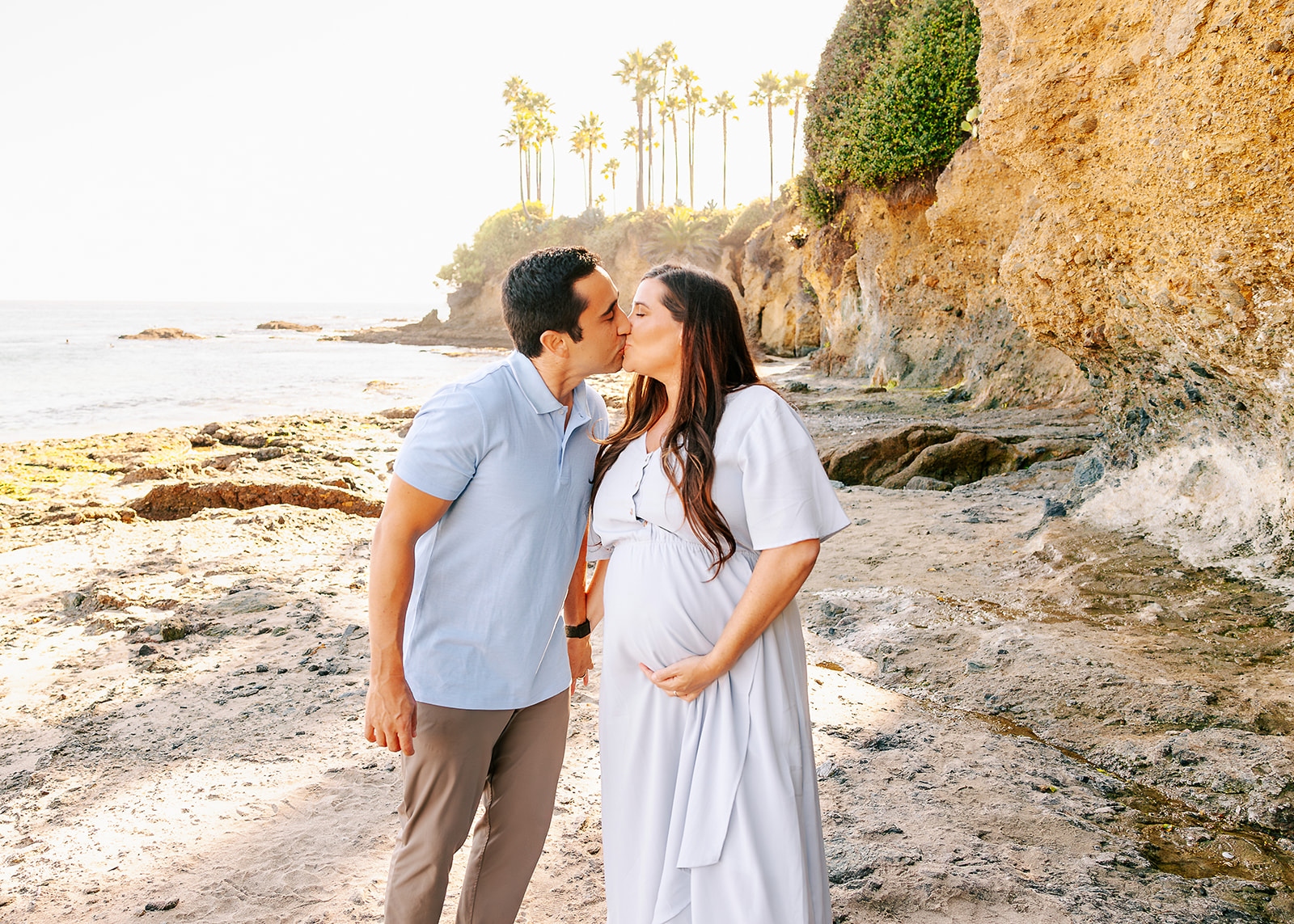 A mom to be kisses her husband while holding hands on a beach at sunset after visiting maternity stores in Boise