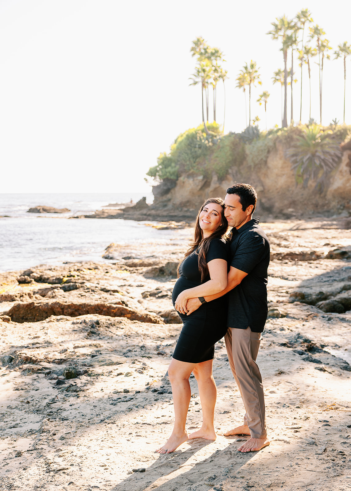 A happy expecting couple in black snuggles and smiles while standing on a beach after visiting maternity stores in Boise