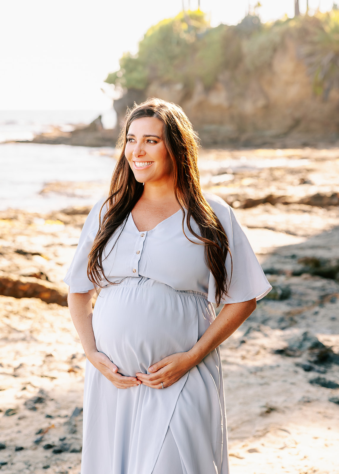 A smiling mother to be walks on a beach with hands on her bump in a blue maternity dress after visiting maternity stores in Boise