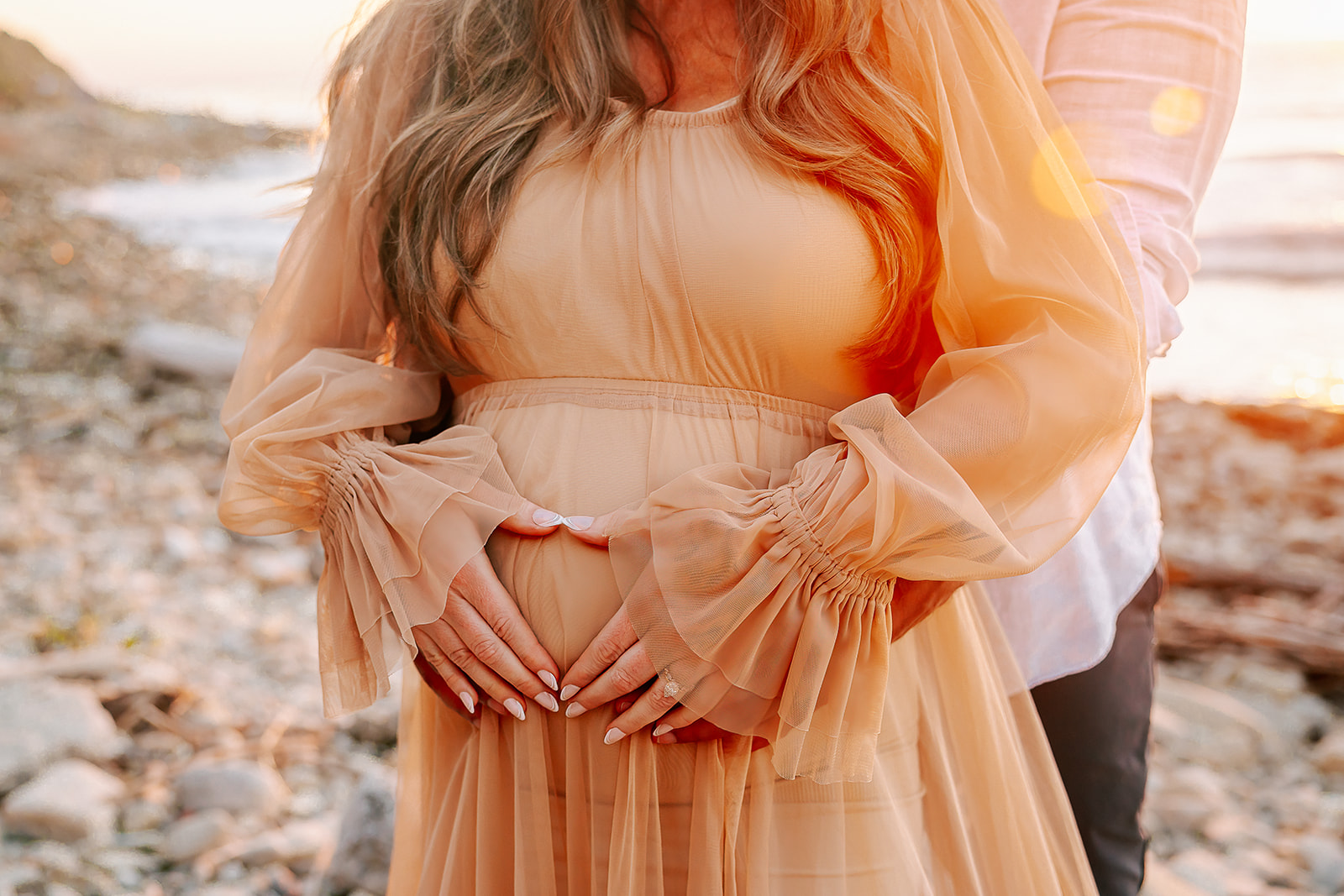 Expecting parents snuggle on a beach with hands in a heart on the bump