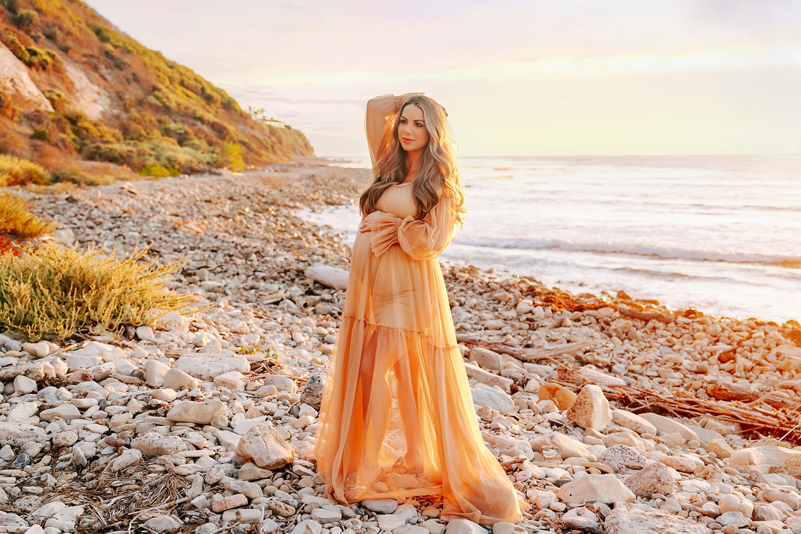 A mom to be walks on a rocky beach at sunset with a hand in her hair in a gold maternity gown