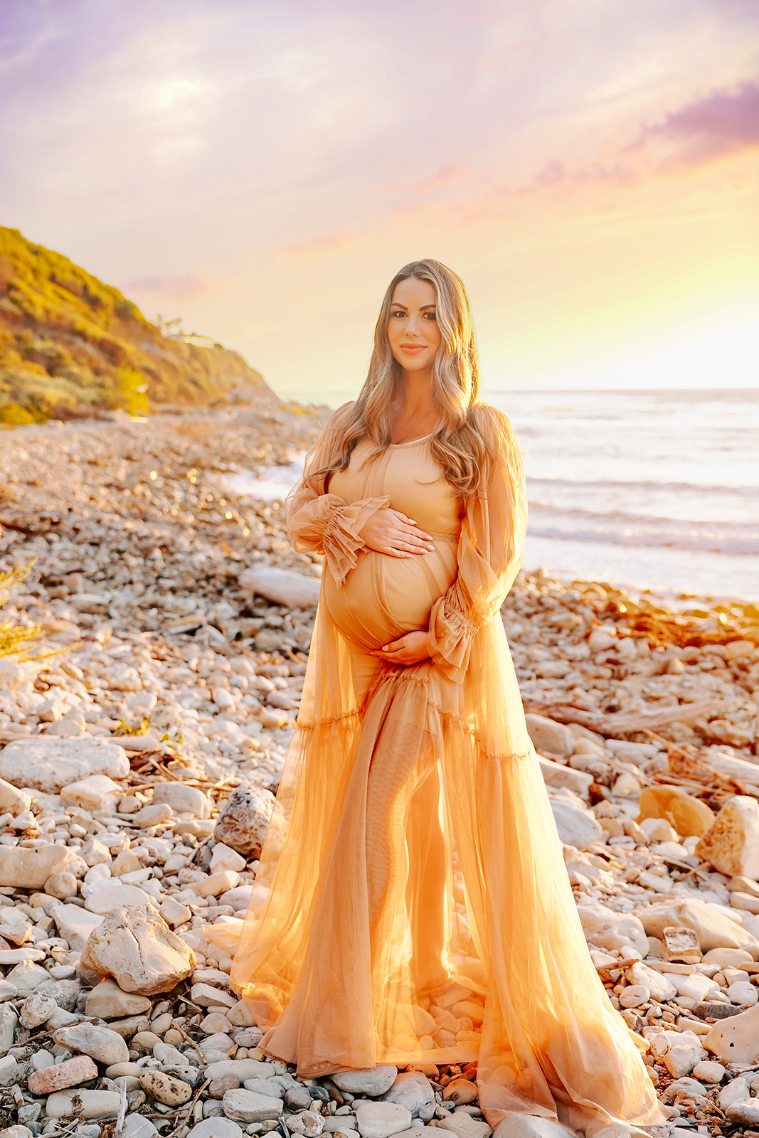 A mother to be in a gold maternity gown stands on a rocky beach at sunset with hands on her bump after some parenting classes in Boise