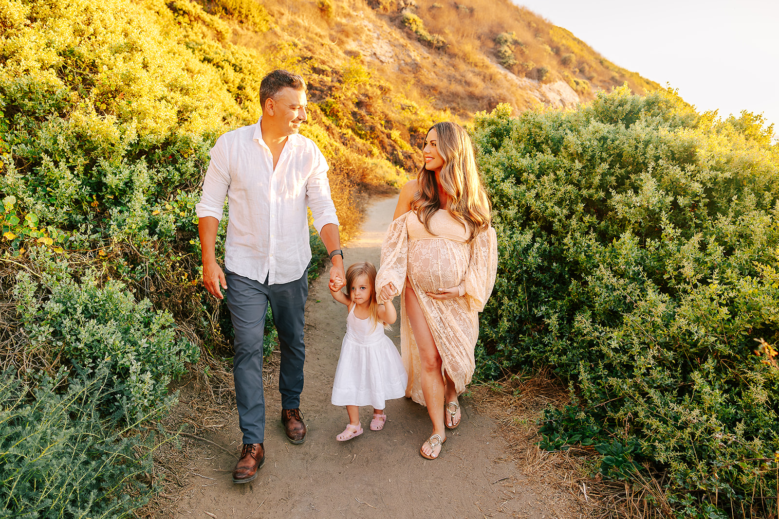 A toddler girl in a white dress walks between pregnant mom and dad holding their hands after some parenting classes in Boise