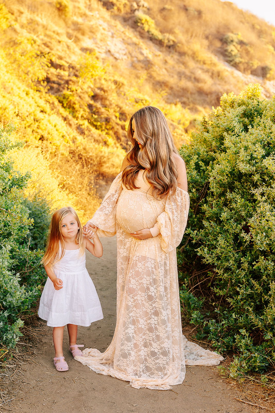 A pregnant mom walks on a trail holding hands with her toddler daughter in a white dress after some parenting classes in Boise