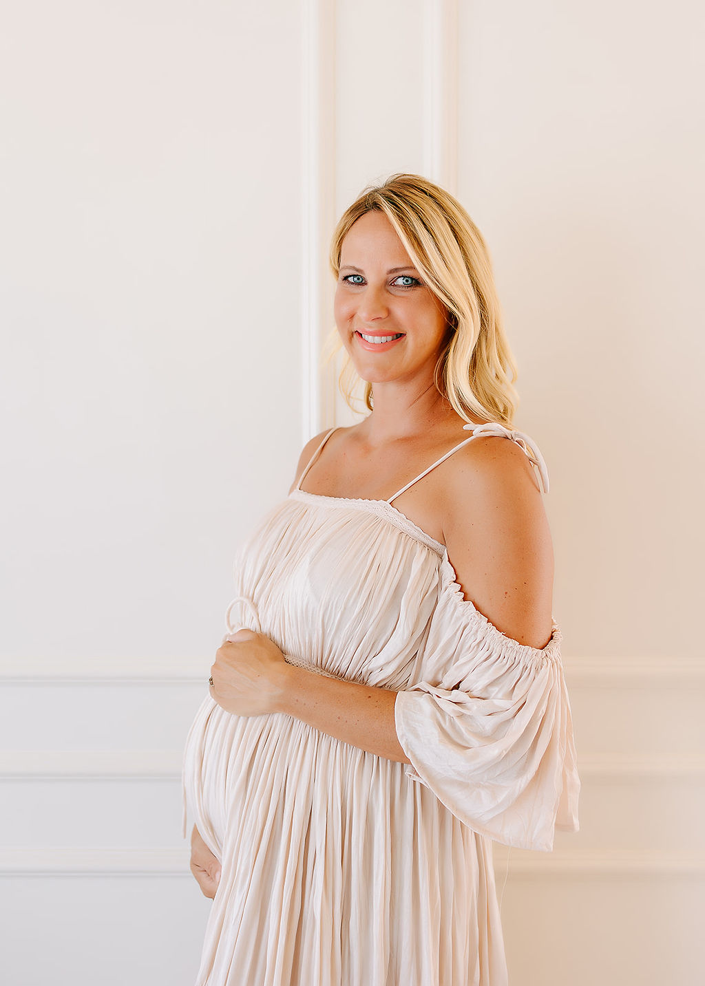 A blonde pregnant woman in a cream maternity dress holds her bump while standing in a white room