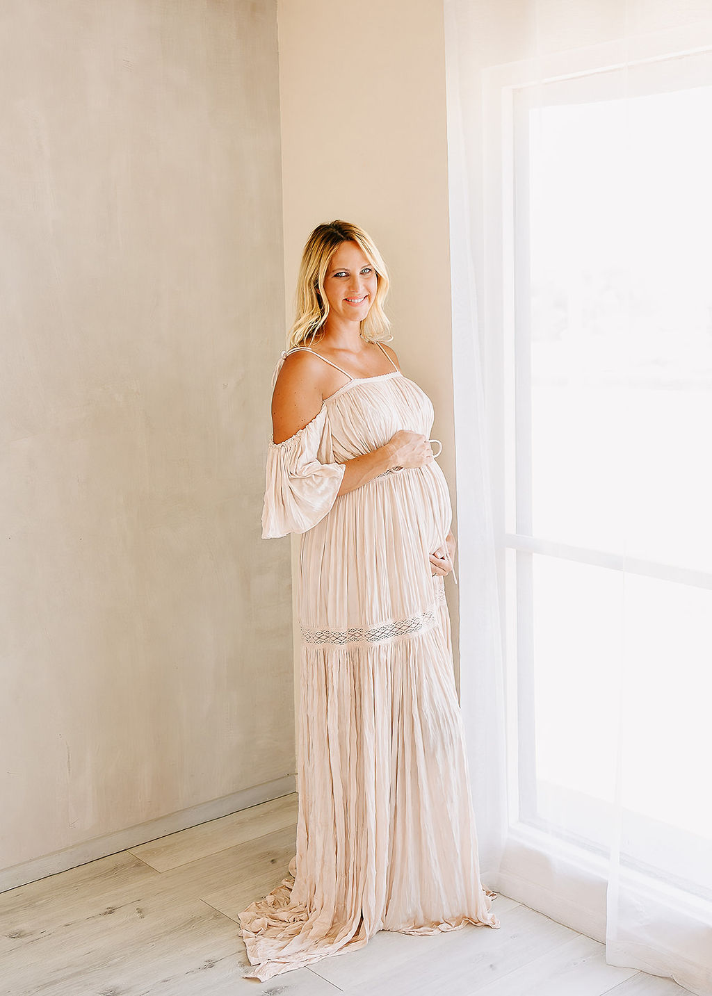 A happy pregnant woman in a beige maternity gown stands in a window holding her bump