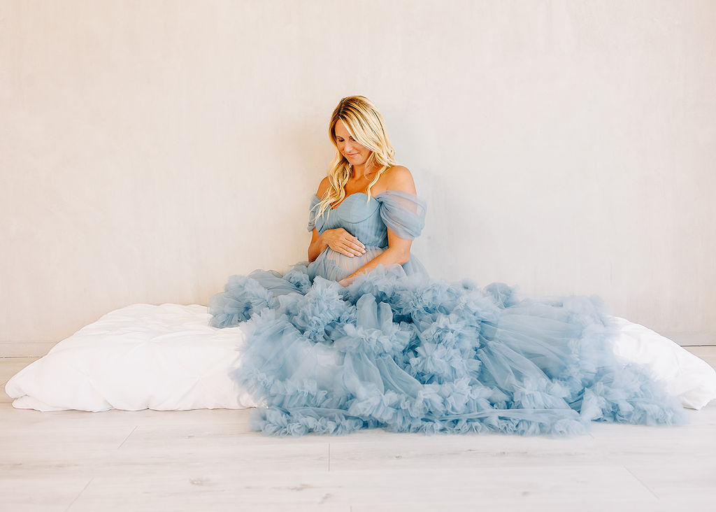 A mother to be in a long blue tule maternity gown sits on a bed against a wall smiling down at her bump before some pelvic floor physical therapy in Boise