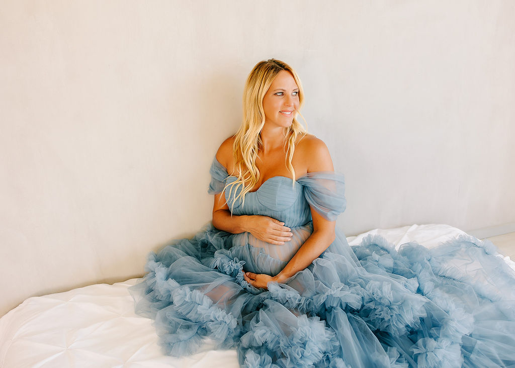 A happy mom to be sits on a bed in a blue tule maternity gown while smiling over her shoulder