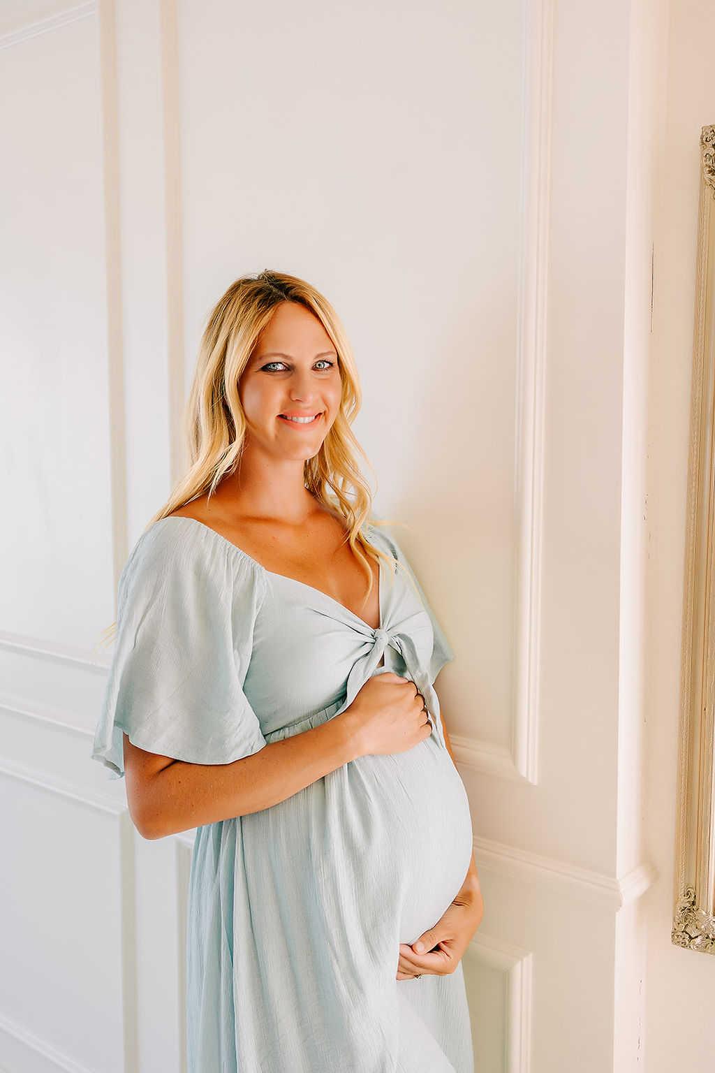 A blonde mother to be in a blue maternity gown holds her bump and leans on a wall before finding pelvic floor physical therapy in Boise