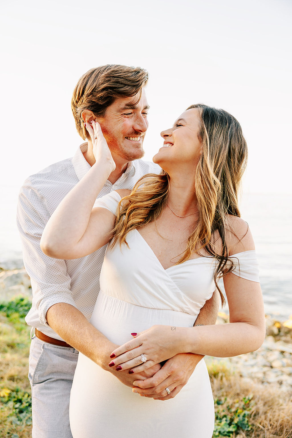 A snuggling couple smiles at each other while standing on a trail by the water at sunset