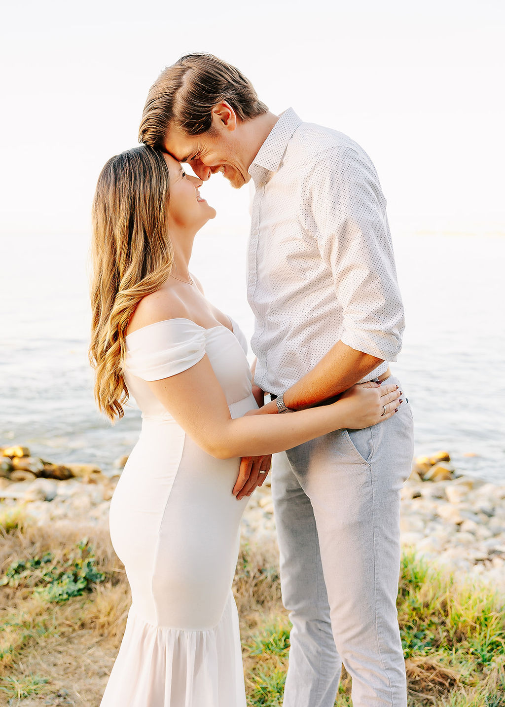 A happy expecting couple giggles and touches foreheads while standing on a beach trail overlook at sunset