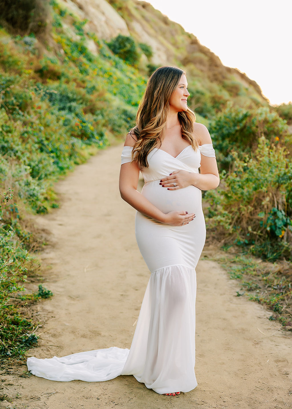 A pregnant woman walks with both hands on her bump in a long white maternity gown while smiling over her shoulder