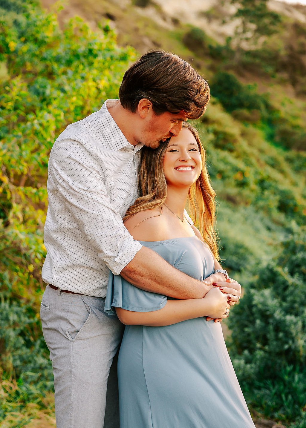 A happy expecting couple snuggles and giggles while standing on a beachside trail at susnet