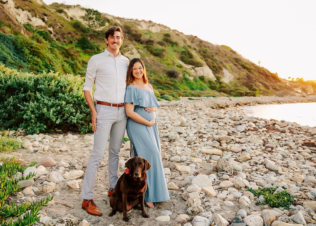 A pregnant woman leans on her tall husband with their brown dog on a rocky beach at sunset after finding placenta encapsulation in Boise