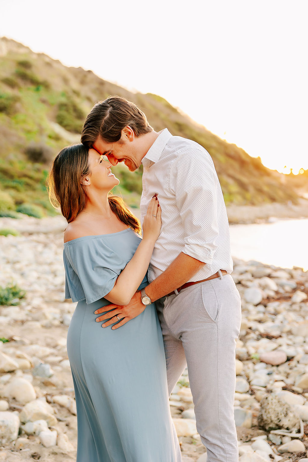 A happy expecting couple in a blue maternity dress and white shirt snuggles foreheads while standing on a rocky beach at sunset after finding placenta encapsulation in Boise