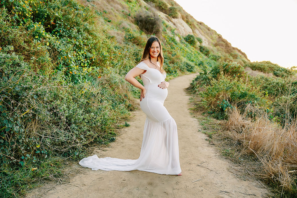 A mother to be stands in a sandy trail in a long white maternity gown with a hand resting on her bump after finding placenta encapsulation in Boise