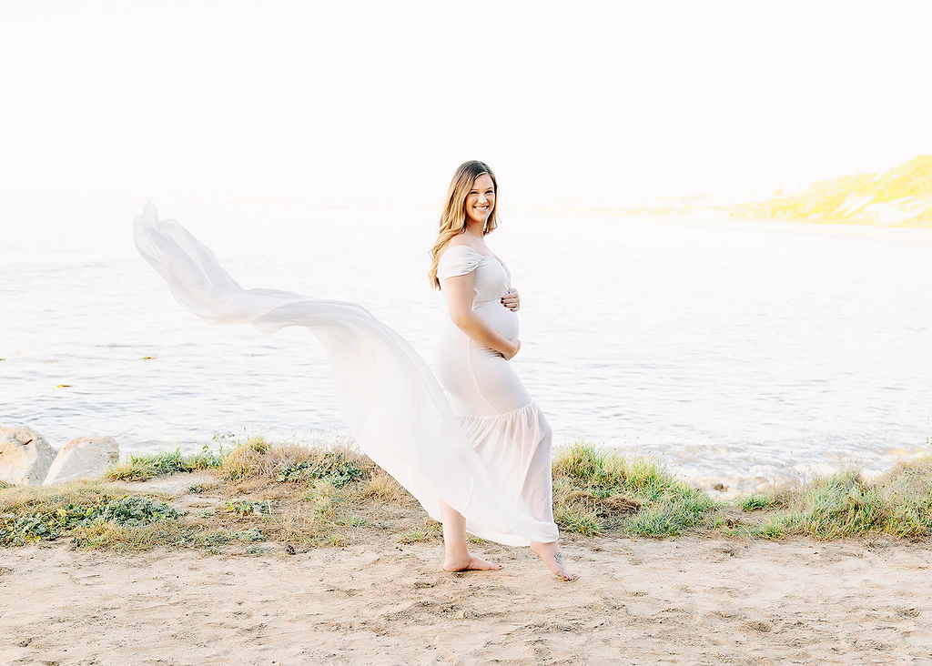 A happy mom to be in a long flowing white maternity gown walks on a beach with hands on bump after meeting for placenta encapsulation in Boise