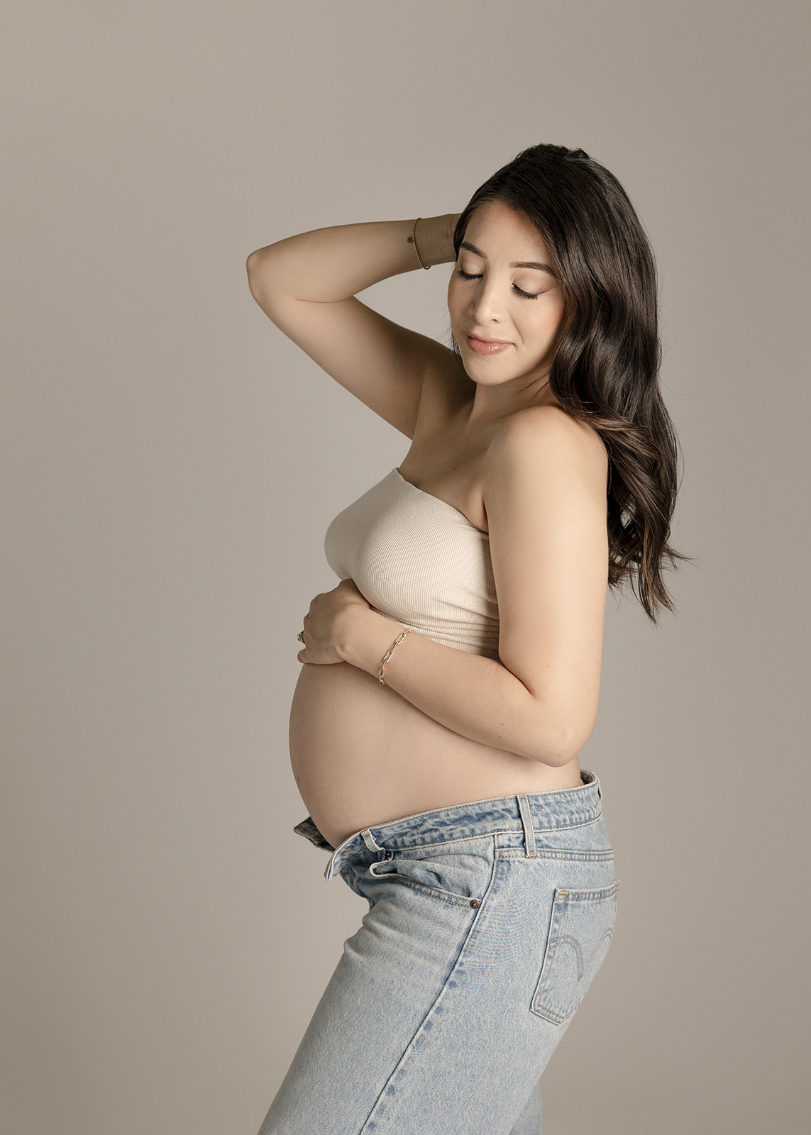 A pregnant woman smiles while resting a hand on the bump and other in her hair in a studio in jeans and tube top after a prenatal chiropractor in Boise