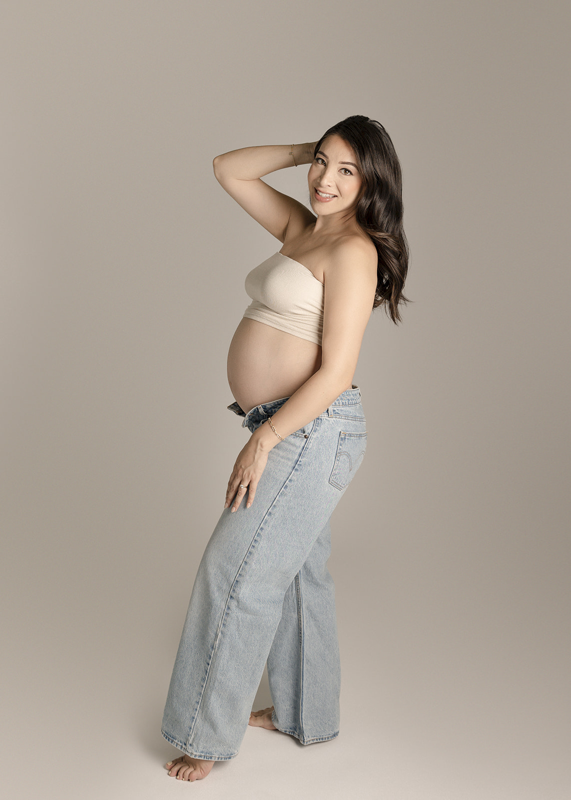 A mother to be in loose open jeans stands in a studio wih a hand in her hair after visiting a prenatal chiropractor in Boise