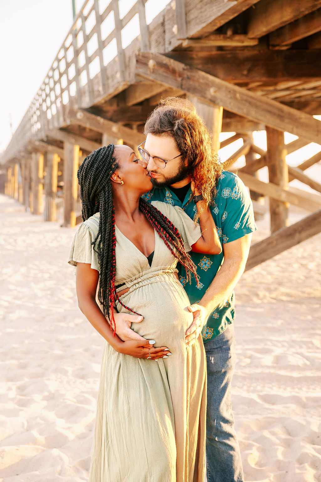 A happy expecting couple holds the bump while kissing on a beach at sunset