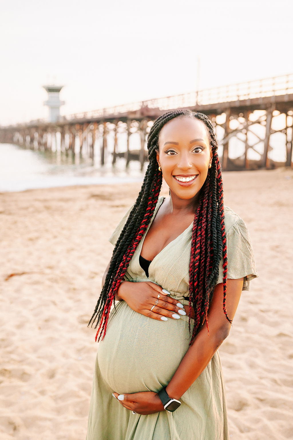 A smiling mom to be in a green maternity gown walking on a beach at sunset by the pier with hands on the bump