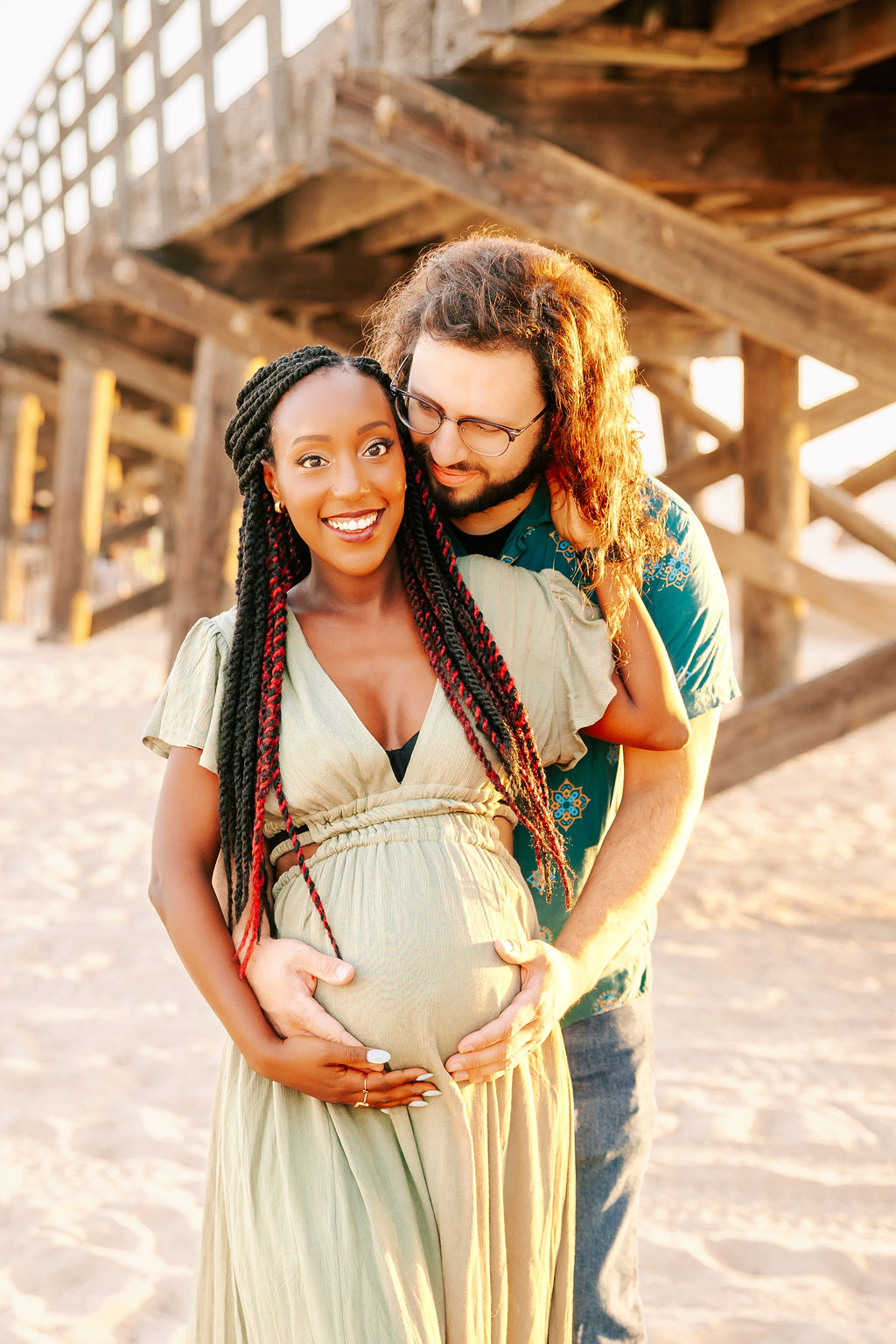 A happy mom to be snuggles with her husband as he hugs her from behind on a beach at sunset under a wooden pier after a prenatal massage in boise