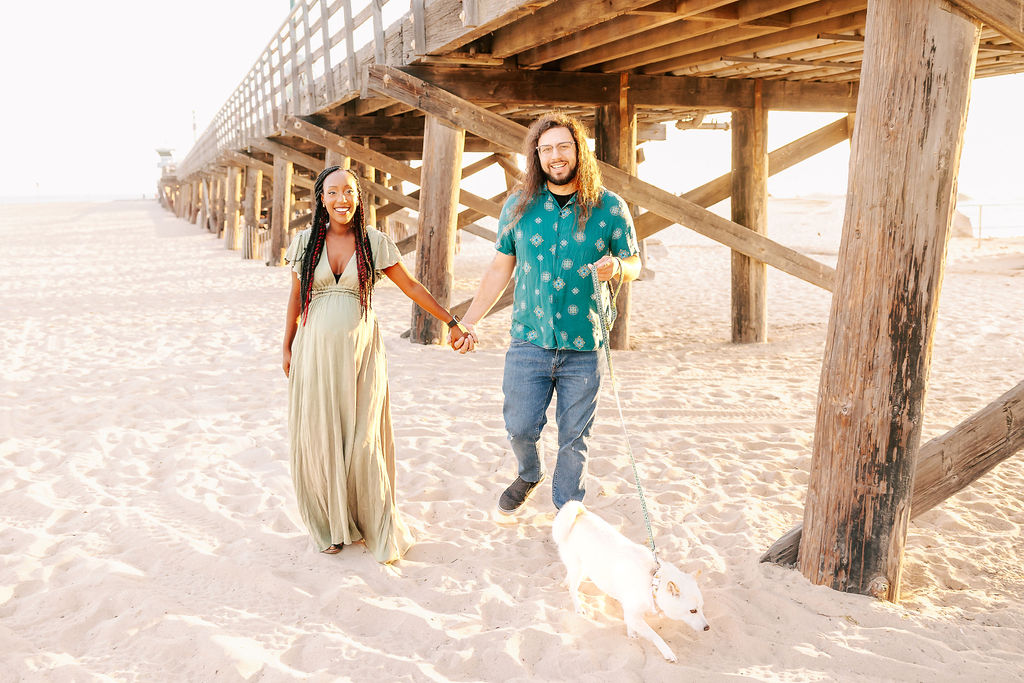 A smiling couple holds hands while walking on a beach at sunset with their white dog under a wooden pier after a prenatal massage in boise